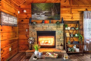 A cozy cabin interior with a fireplace, wooden walls, a painting of a forest stream, and a dining table with food and drinks. - Romantic - Firepit - Hot tub - Pet Friendly (Sevierville)