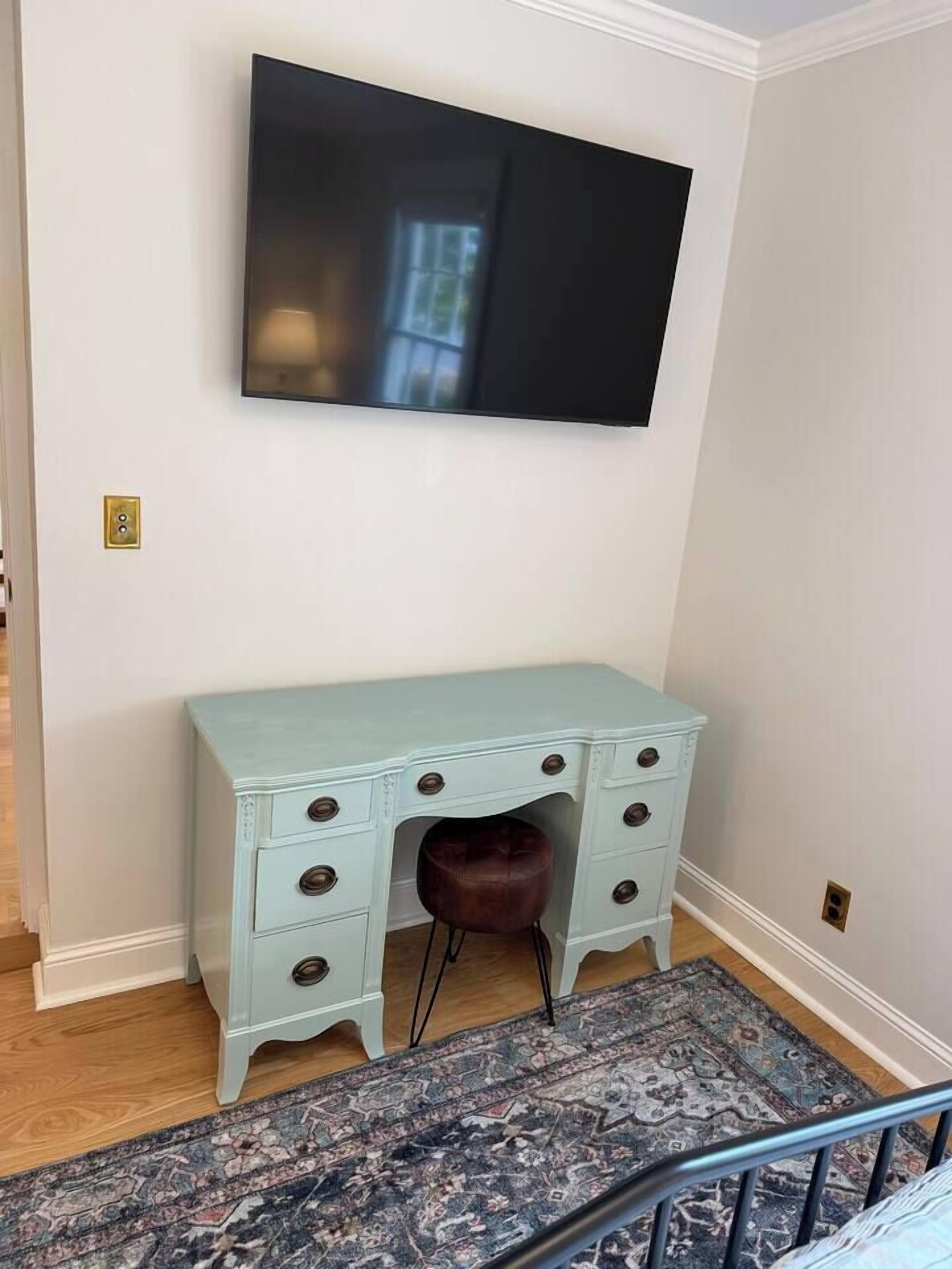 A room with a light blue desk, a brown chair, a patterned rug, and a flat-screen TV mounted on the wall.