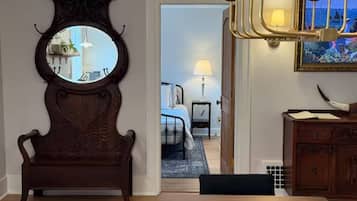 A room with a wooden bench, a round mirror, a door leading to another room with a bed and a nightstand.