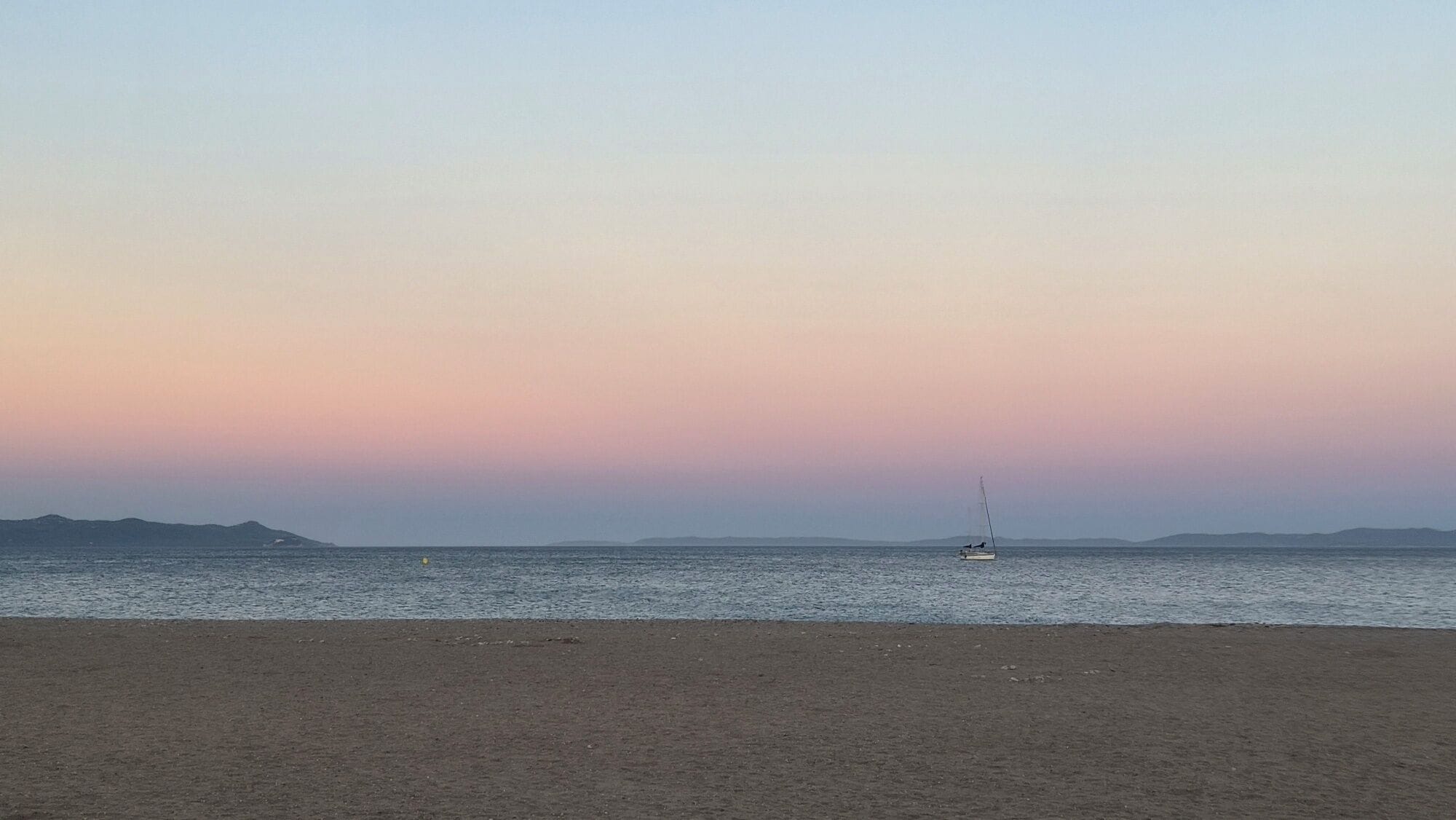 A beach with a sailboat on calm waters, distant mountains, and a clear sky.