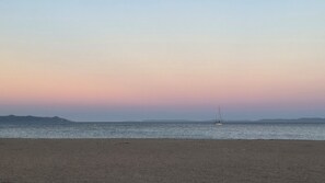 Una platja amb un veler sobre aigües tranquil·les, muntanyes llunyanes i un cel serè.