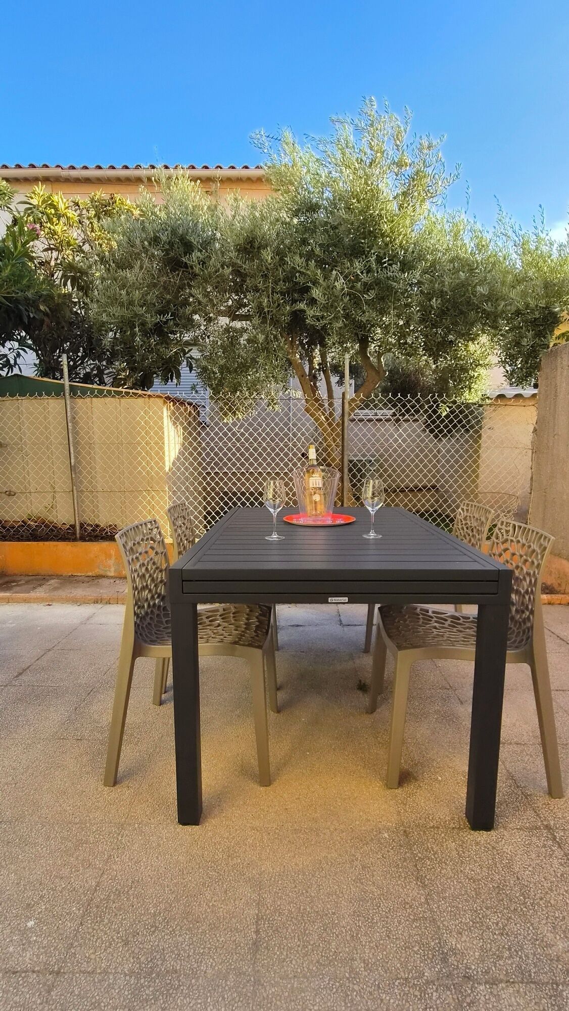 An outdoor patio with a table and chairs, a bottle of wine, and a fence.