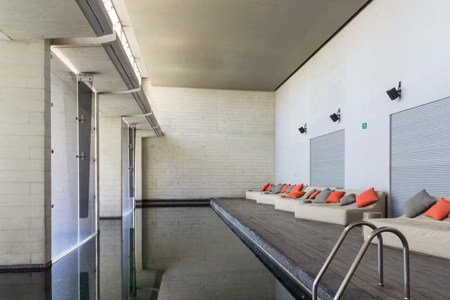 A modern indoor pool area with a long, narrow pool, lounge chairs with cushions, and a metal ladder leading to the pool.