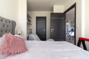 Een moderne slaapkamer met een gestoffeerd hoofdeinde, een groot bed met een zacht kussen, een bureaustoel, een kledingkast en een eethoek zichtbaar door een open deur.