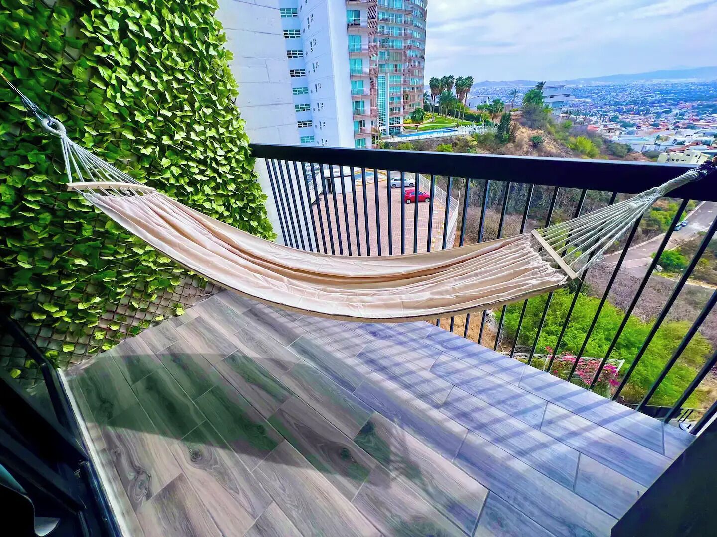 A hammock on a balcony with a view of a cityscape and a green wall.