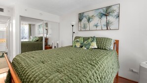 A bedroom with a large bed, a mirror, a painting of palm trees, and a bedside lamp. - Waikiki Beach Airbnb | Pool, Parking,Walk to Beach (Honolulu)
