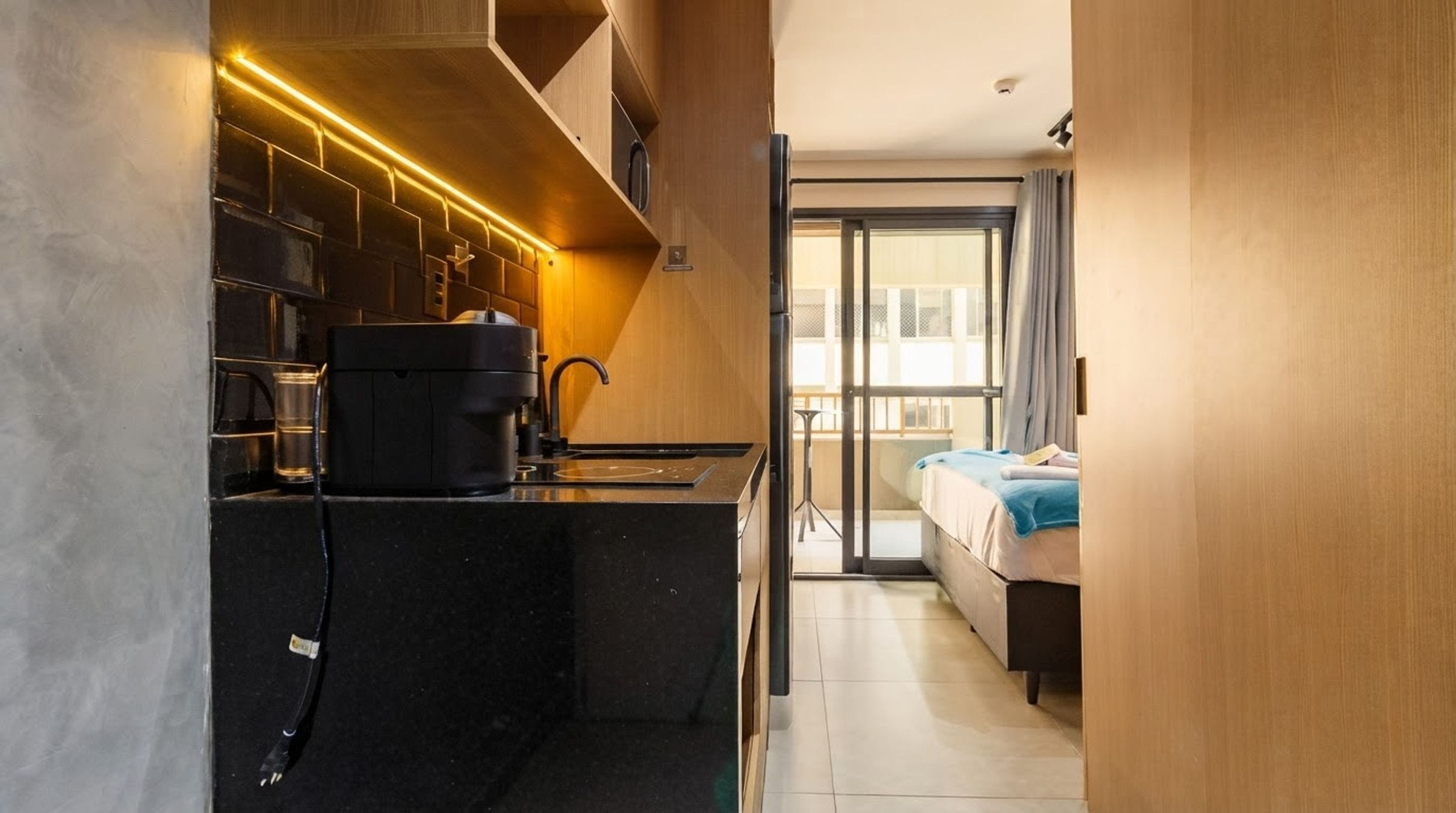A compact kitchen with a coffee machine, a sink, and a stove. There is a sliding glass door leading to a balcony with a bed.