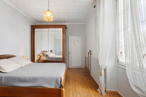 Un dormitorio con cama de madera, un espejo, una lámpara colgante y una ventana con cortinas translúcidas.