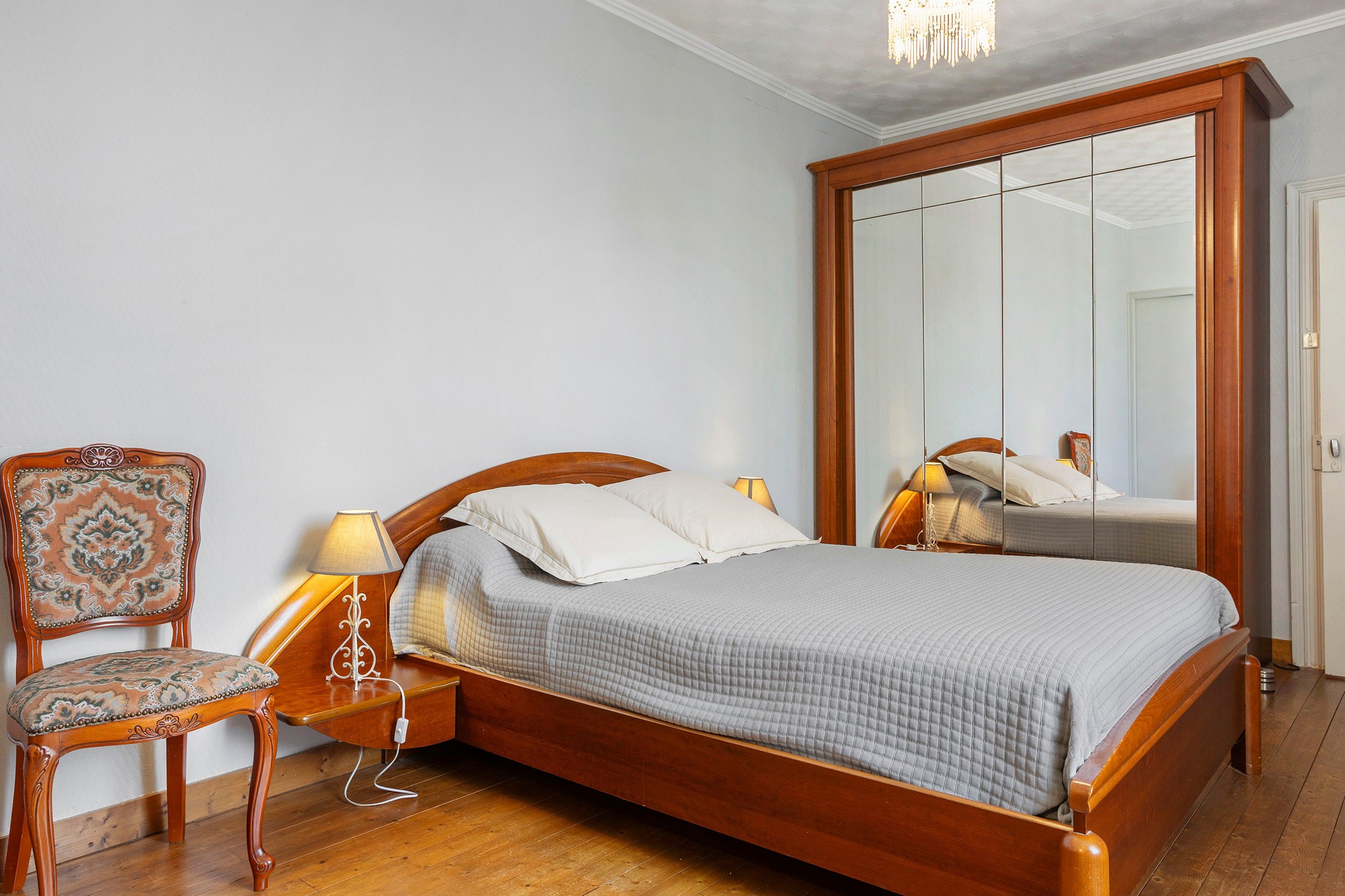 A bedroom with a wooden bed, a chair, a nightstand, and a large mirror.