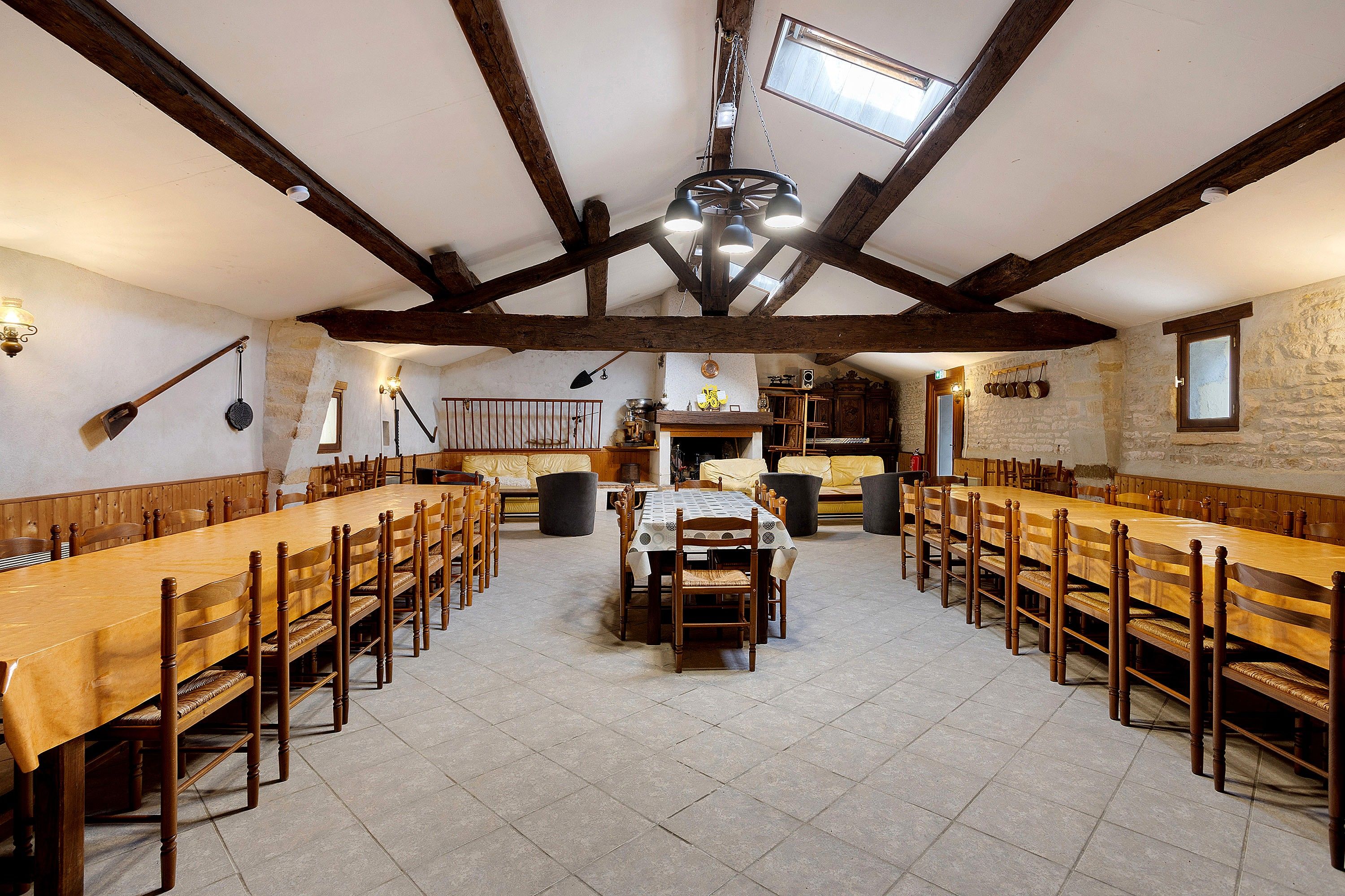 A large dining hall with high ceilings and exposed wooden beams, featuring a central table surrounded by long wooden tables and chairs.