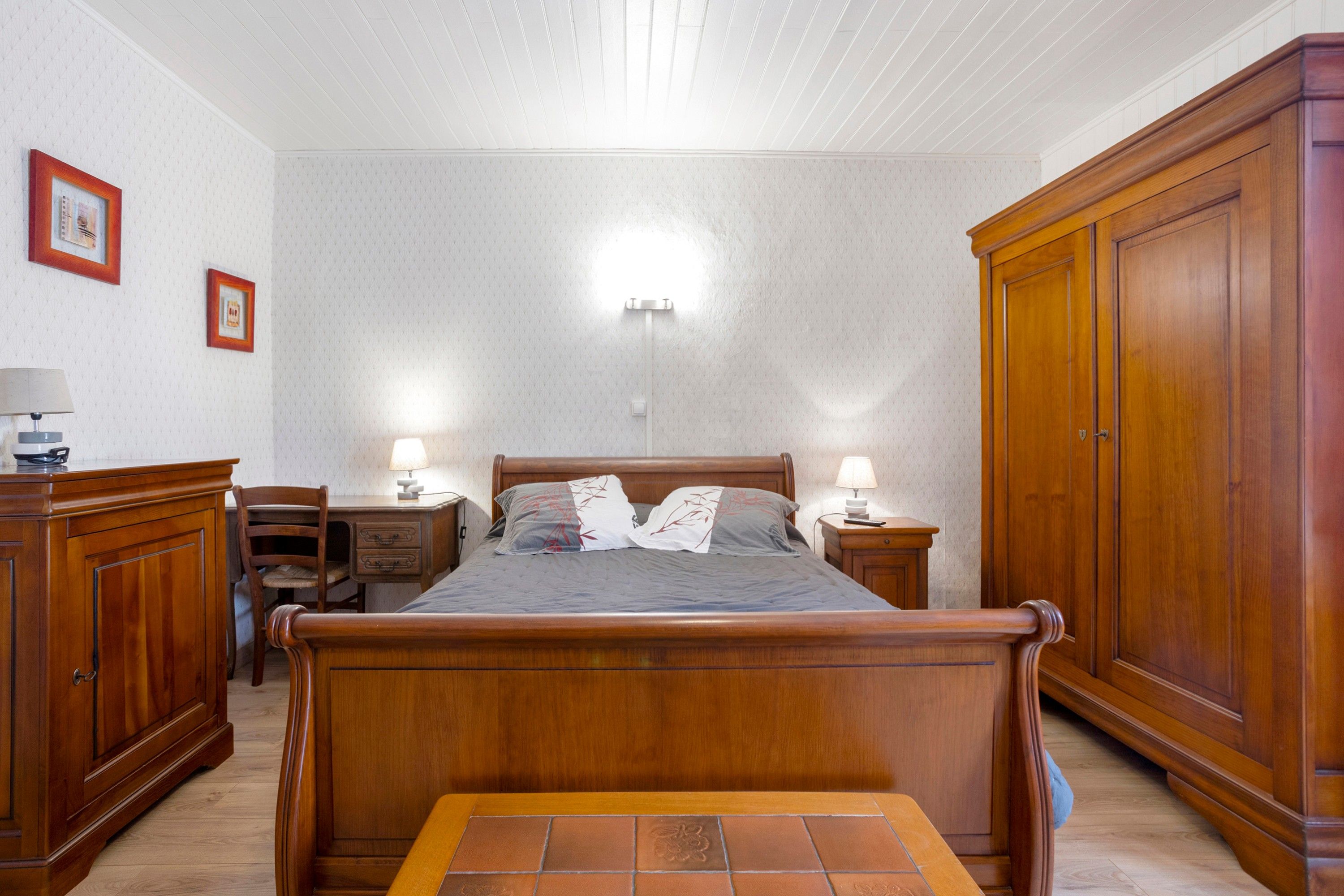 A bedroom with a wooden bed, bedside lamps, a nightstand, a wardrobe, and a desk with a chair.