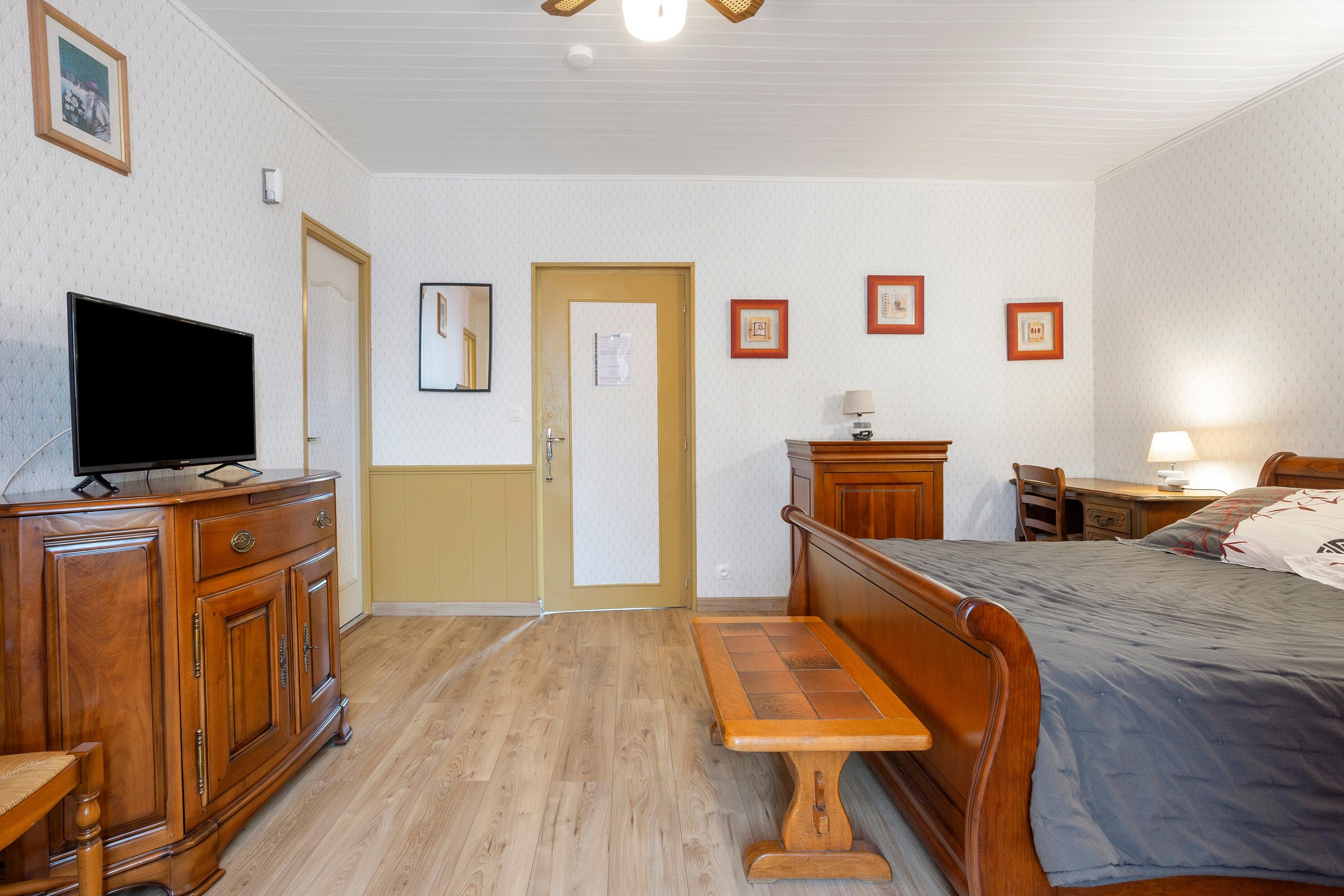 Habitación de hotel con una cama de madera, un televisor en un aparador, un escritorio con lámpara y una puerta con espejo.