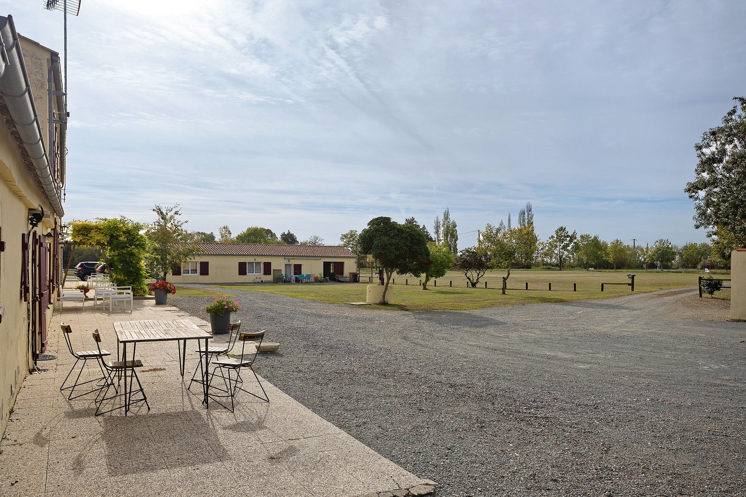 A gravel courtyard with a table and chairs, a building with a tiled roof, and a grassy field.