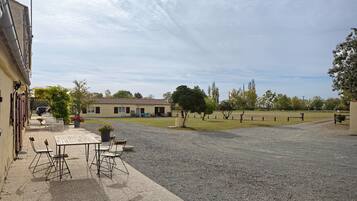 A gravel courtyard with a table and chairs, a building with a tiled roof, and a grassy field.