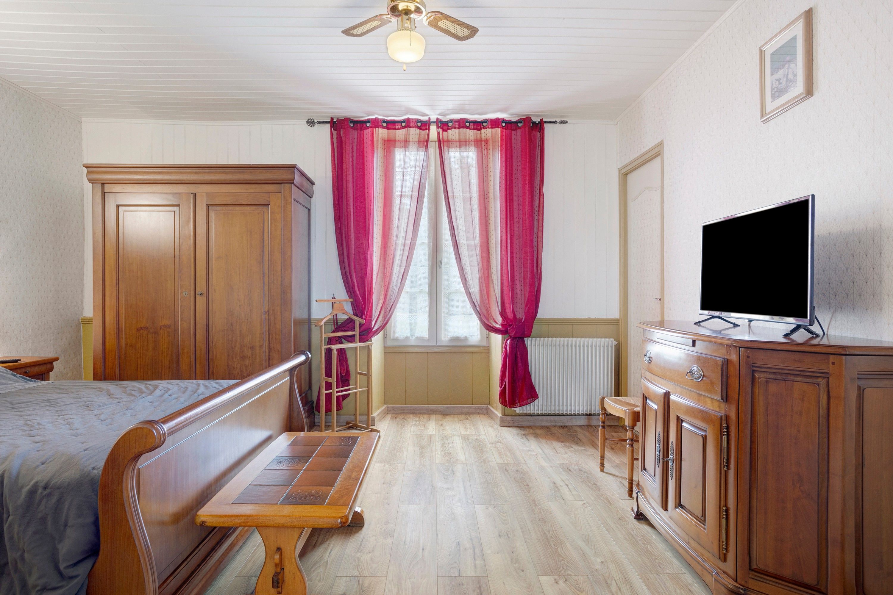 Une chambre à coucher avec un lit, une commode en bois, un téléviseur et une fenêtre avec des rideaux.