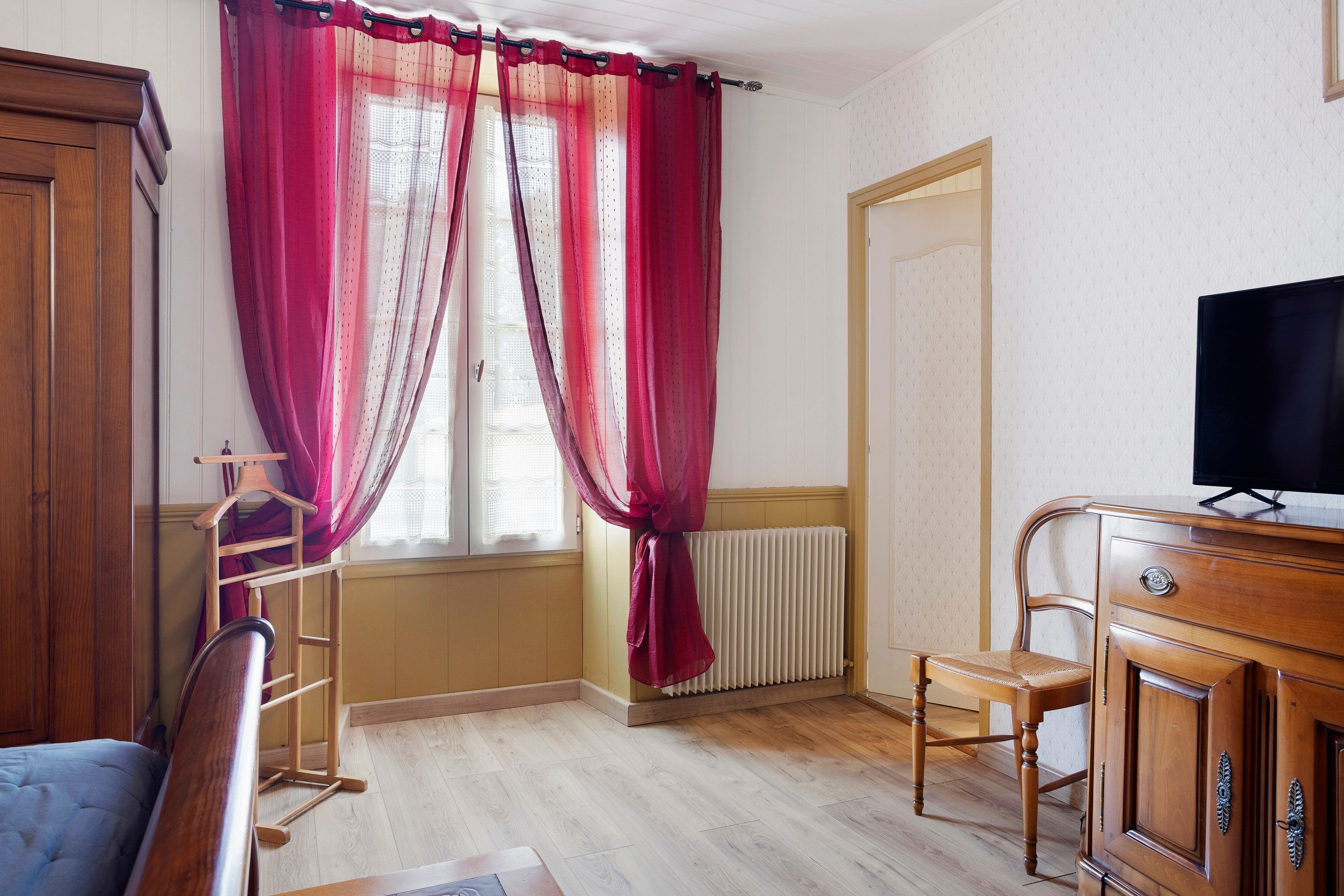 Une pièce avec une armoire en bois, un téléviseur posé sur un meuble en bois, un radiateur et une fenêtre avec des rideaux rouges.