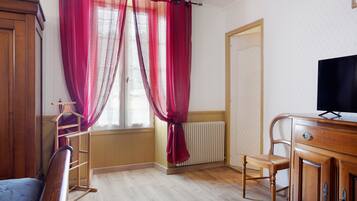 Une pièce avec une armoire en bois, un téléviseur posé sur un meuble en bois, un radiateur et une fenêtre avec des rideaux rouges.