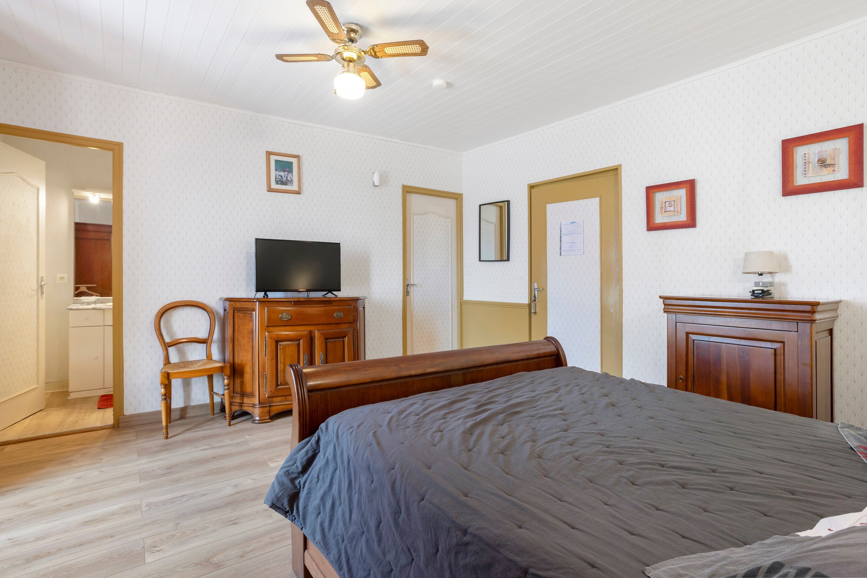 Een slaapkamer met een bed, een televisie op een houten dressoir, een stoel en een plafondventilator.