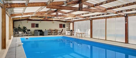 A swimming pool with a wooden ceiling and walls, surrounded by lounge chairs and a table.