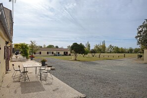 A gravel courtyard with a table and chairs, a building with a tiled roof, and a grassy field. - Le Paradis - Chambre d'hôtes Val pour 4 pers (Chaillé-les-Marais)