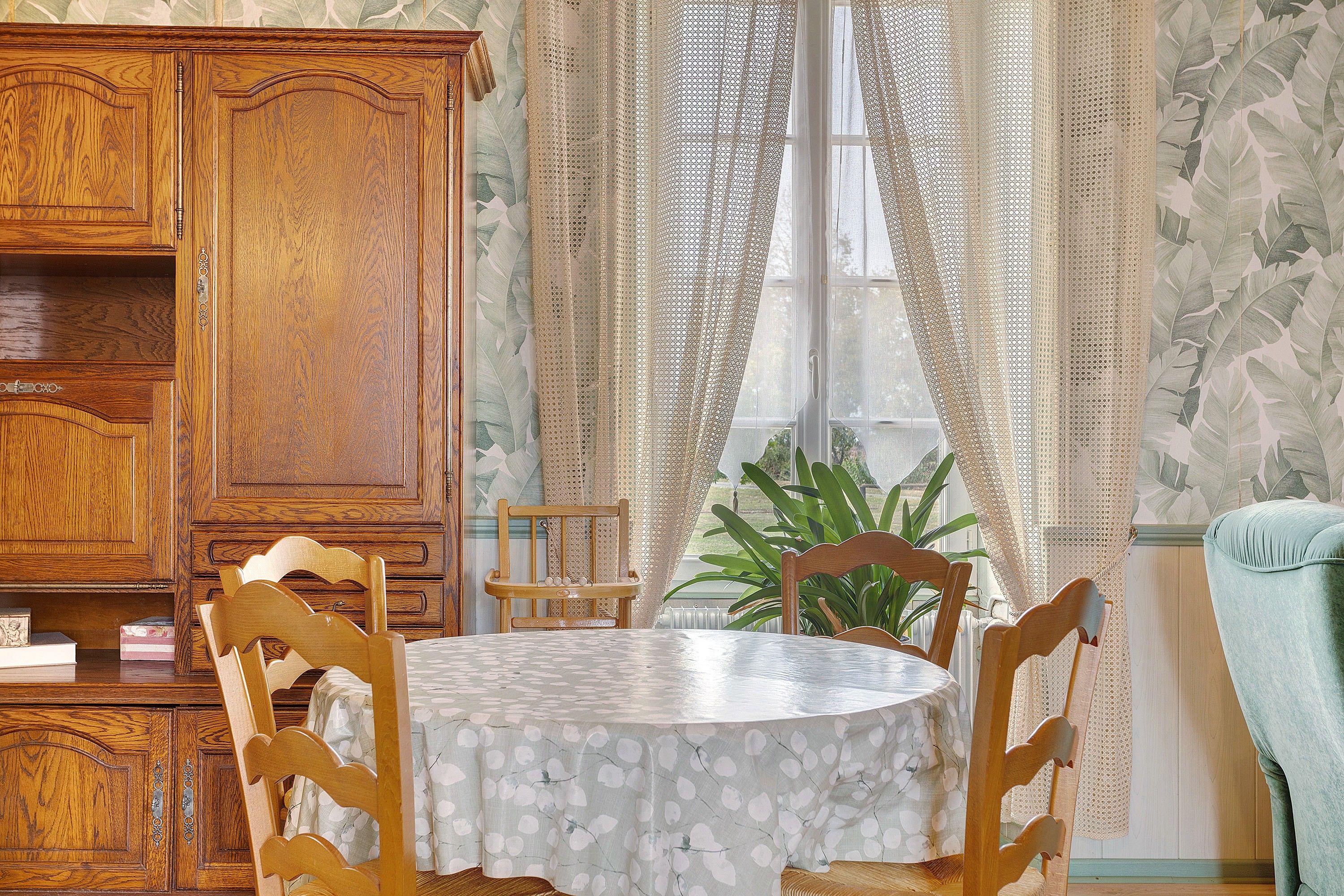 Uma sala de jantar com mesa redonda, cadeiras de madeira, um armário e janela com cortinas.