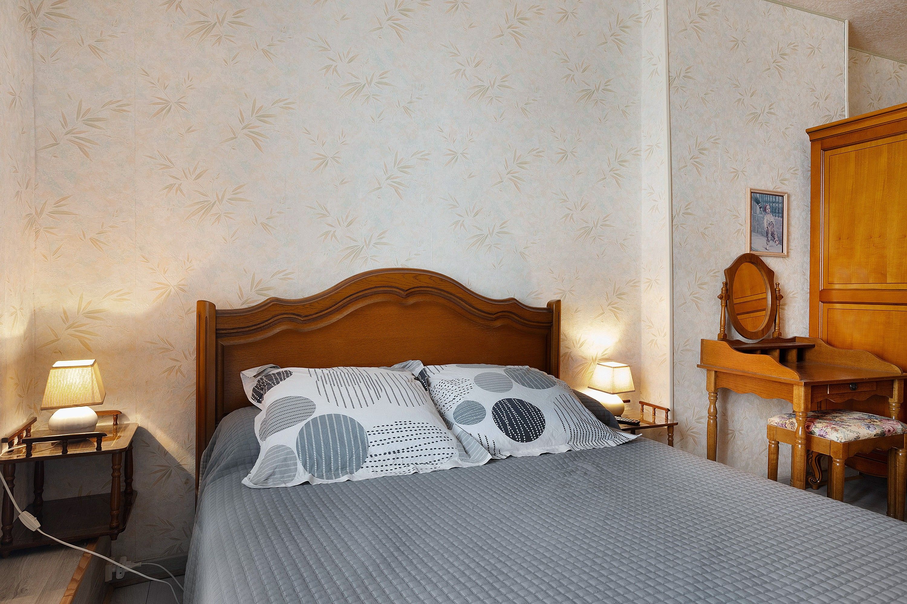 A neatly made bed with a patterned bedspread and pillows, flanked by bedside tables with lamps, a wooden nightstand, and a wooden chair with a cushion.