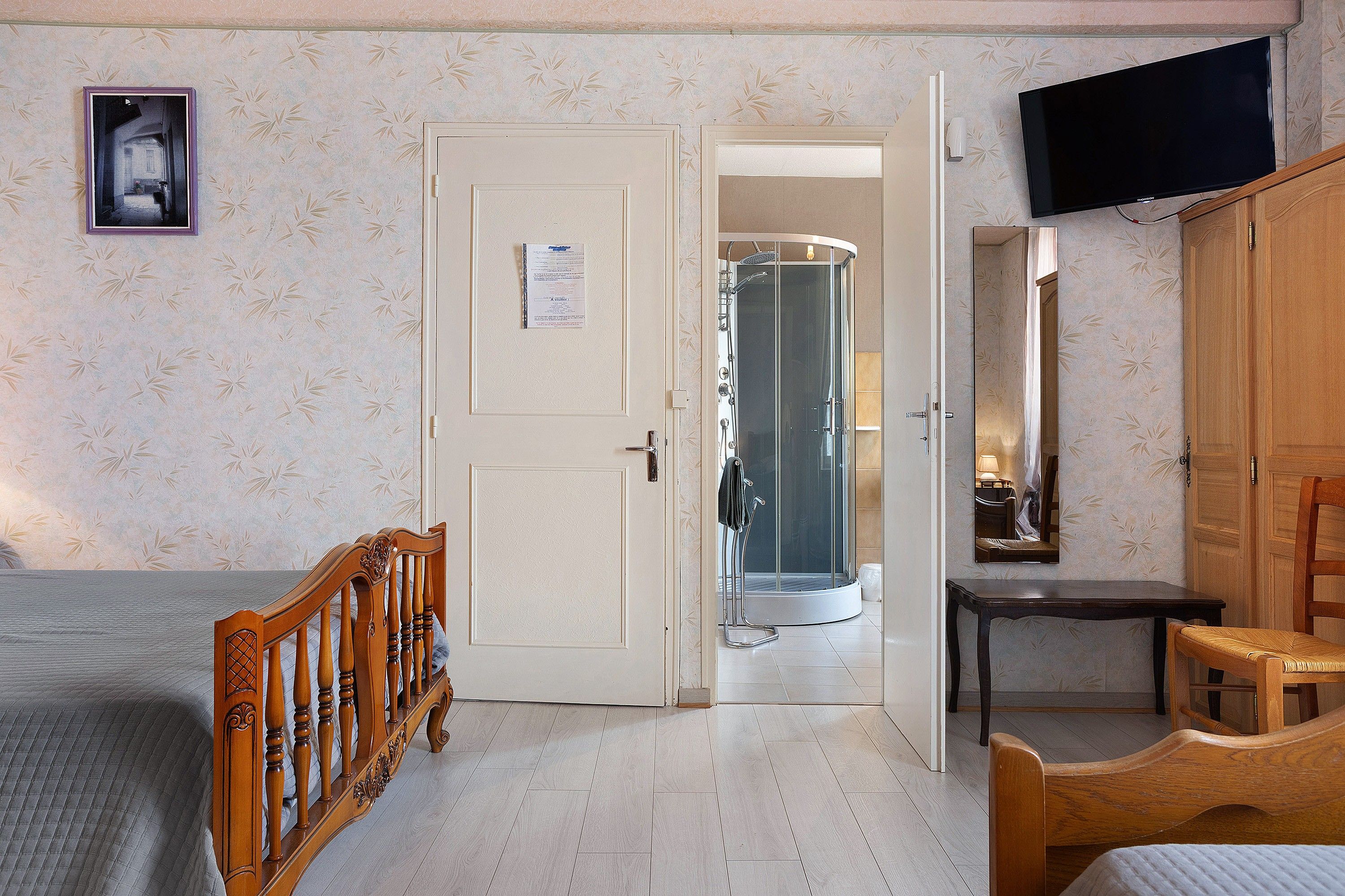 Ein Hotelzimmer mit einem Bett, einem Holzstuhl, einem kleinen Tisch, einem Fernseher und einem Duschbereich.