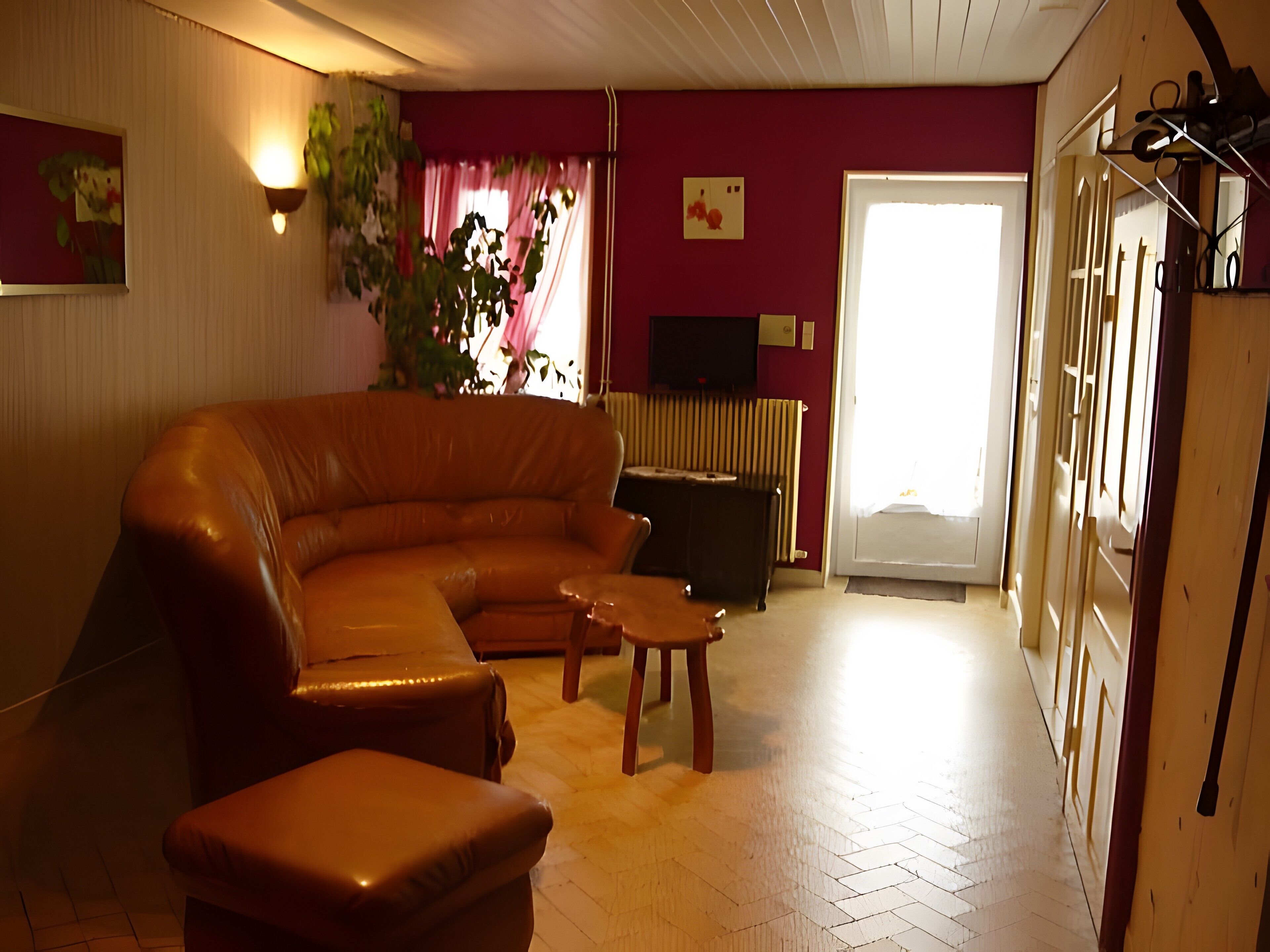 Sala de estar con un sofá de cuero, una mesa de centro de madera y una mesita auxiliar. Hay un televisor sobre un mueble, una planta y una puerta que da a otra habitación.