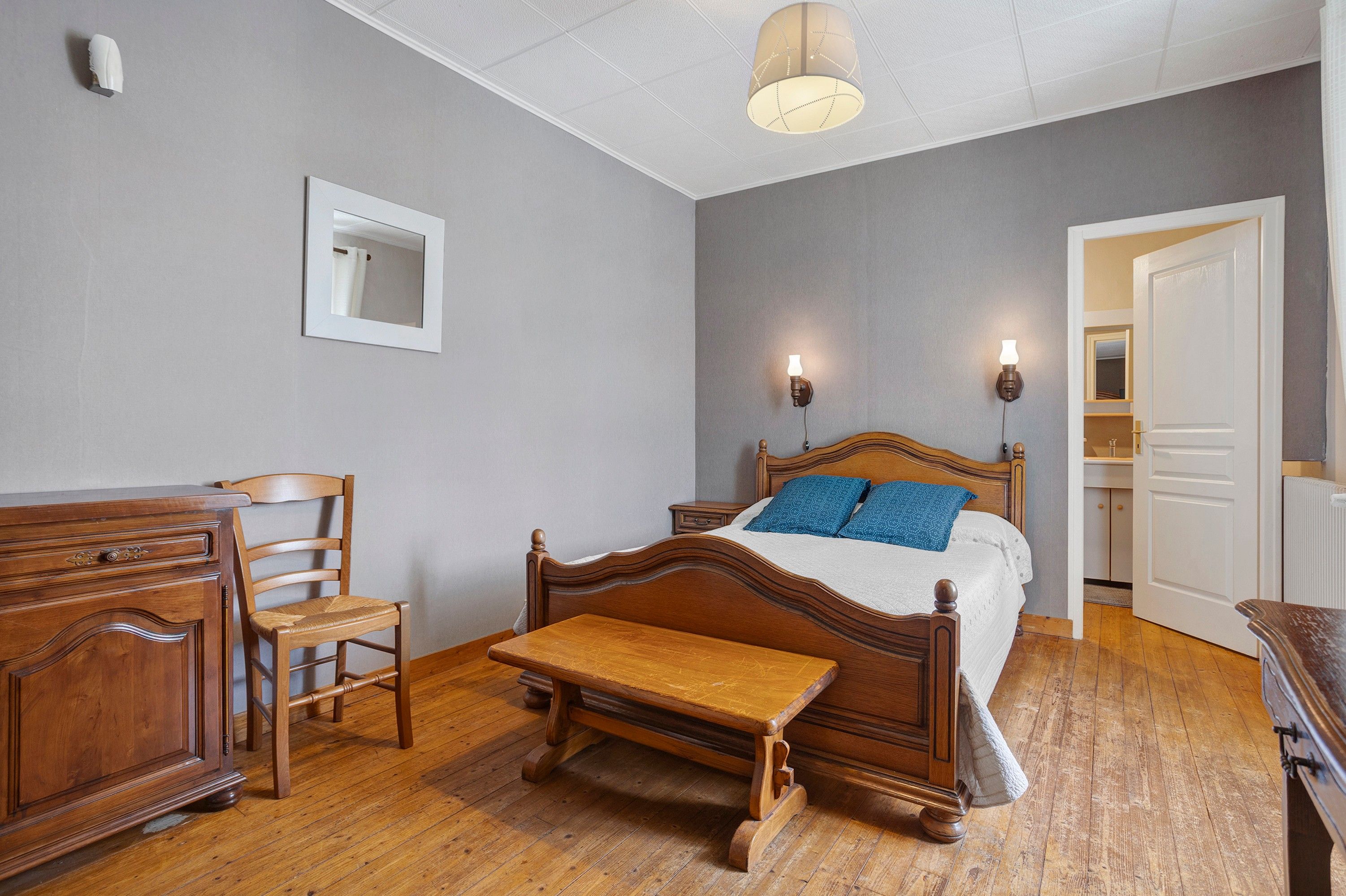 Ein Schlafzimmer mit einem hölzernen Bett, einem Nachttisch, einem Stuhl, einer Holzbank, einem Spiegel und einer Tür zu einem Badezimmer.