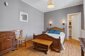 Un dormitorio con cama de madera, una mesita de noche, una silla, un banco de madera, un espejo y una puerta que da a un baño.