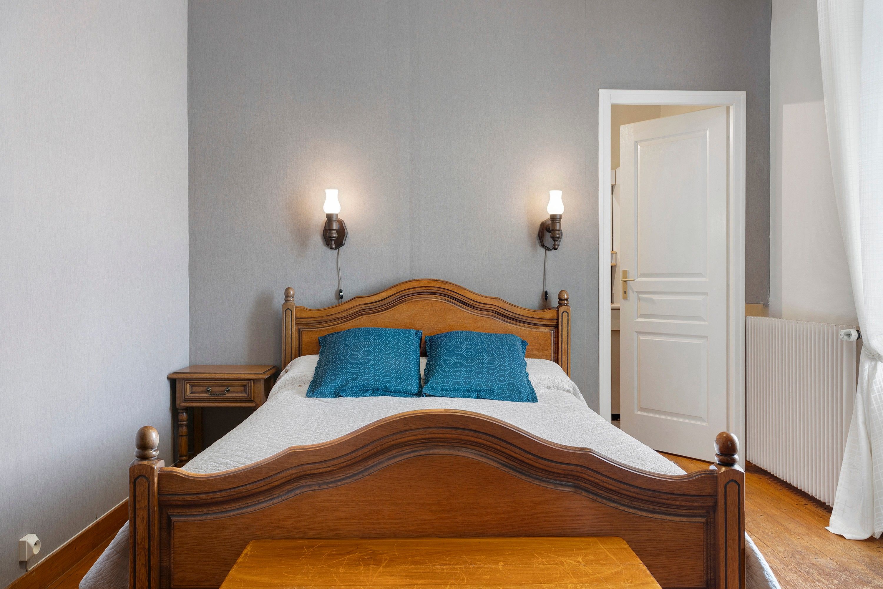 Une chambre à coucher avec un lit en bois, deux lampes fixées au mur, une table de chevet et une porte.