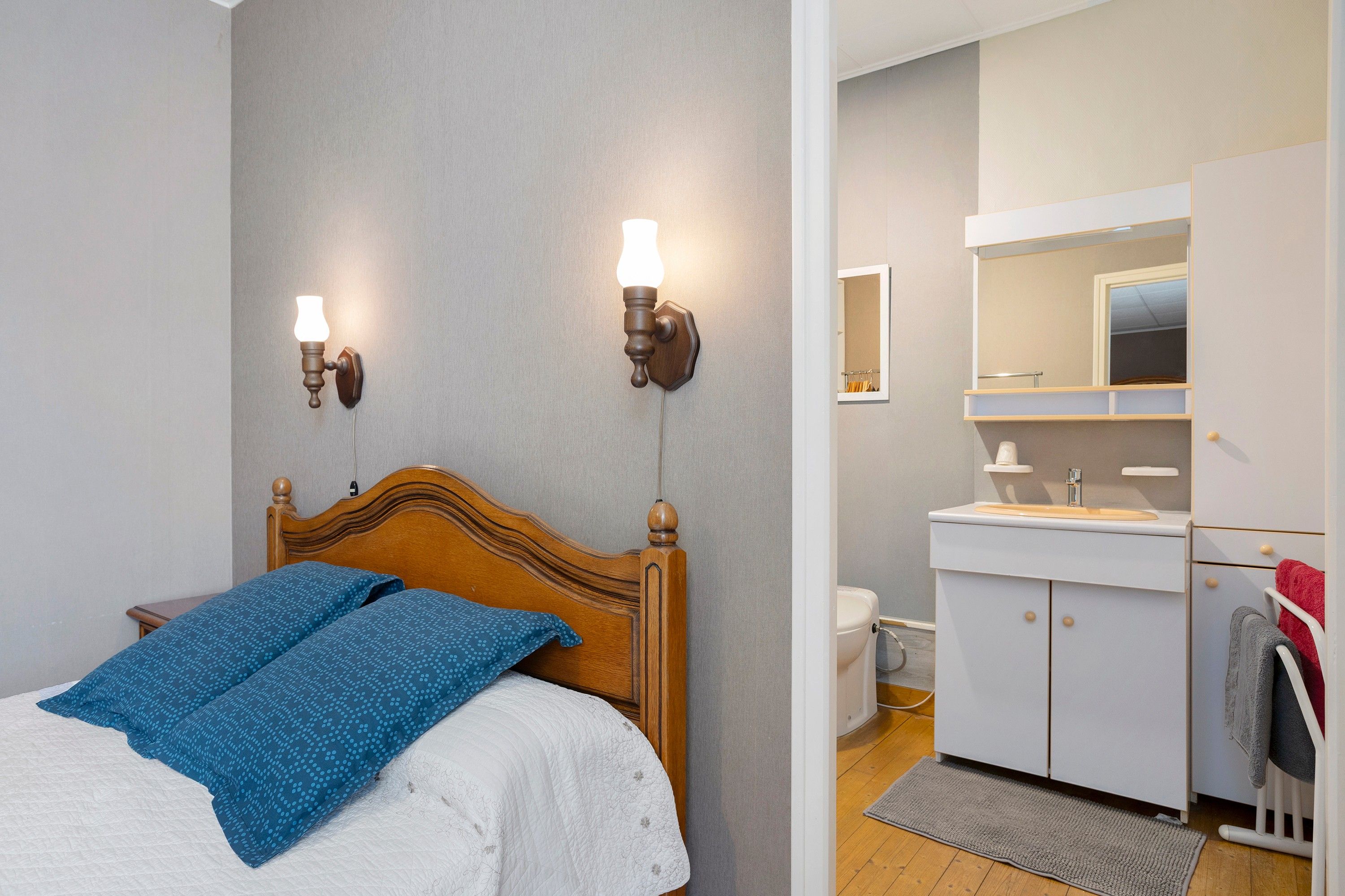 Une chambre à coucher avec une tête de lit en bois, deux lampes fixées au mur, un lit avec une literie bleue et blanche, et une salle de bain avec un lavabo et un miroir blancs.