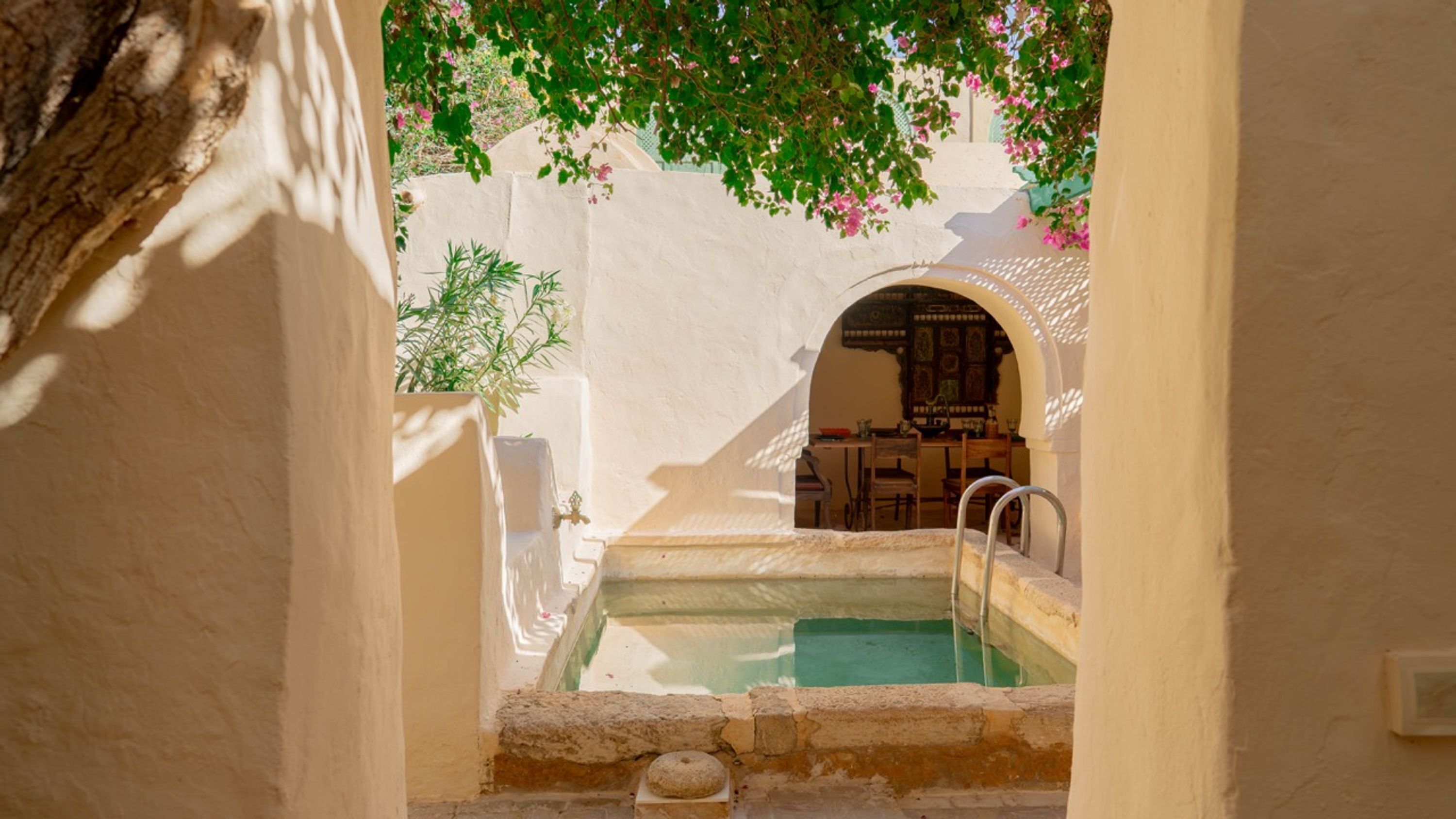 A pool area with a stone bench, a table, and chairs, surrounded by walls and a tree.