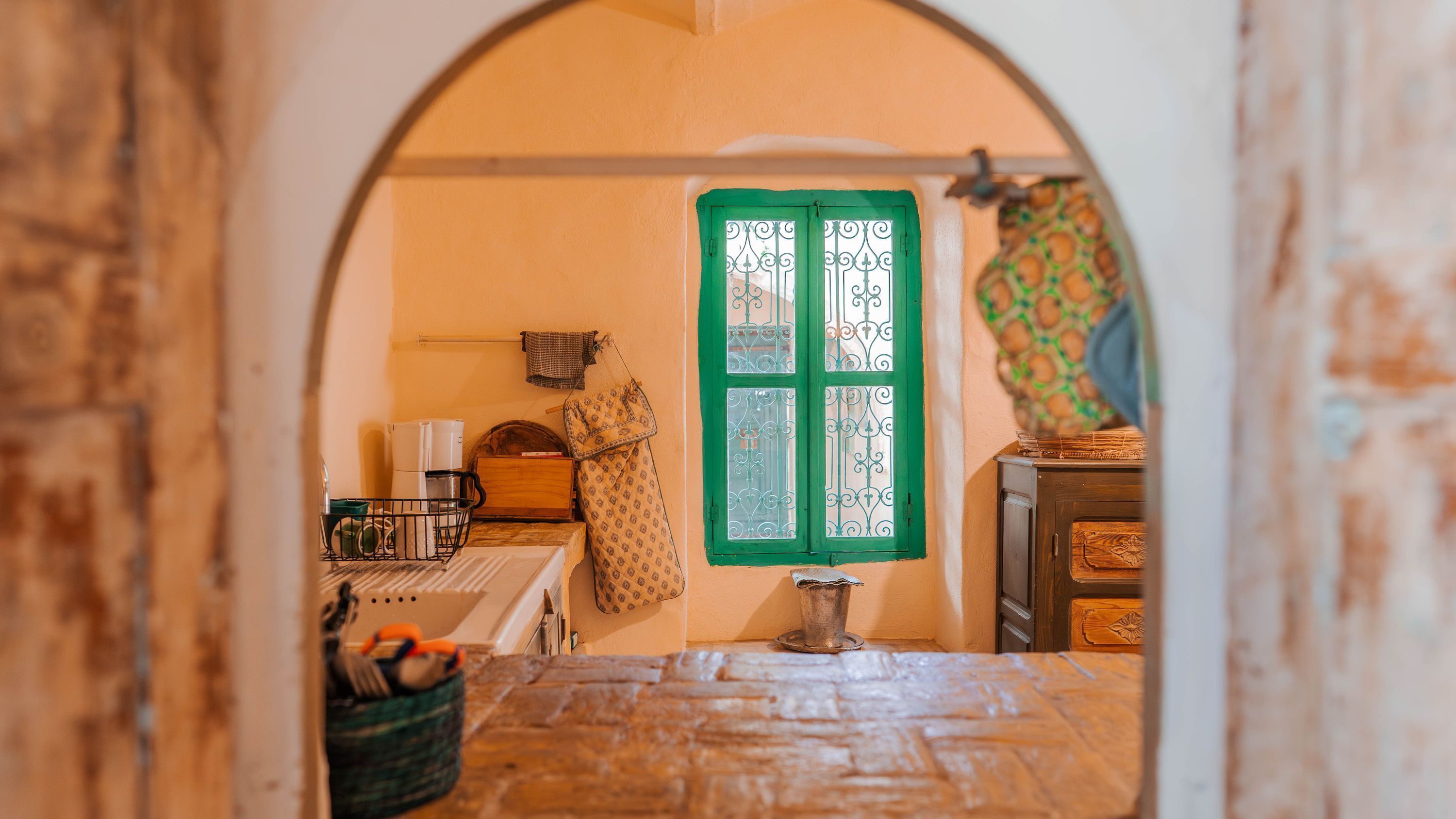 A narrow hallway with a green-framed window, a small kitchen area with a refrigerator, and a patterned wall.