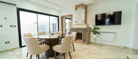 A modern living room with a fireplace, a dining table with chairs, a flat-screen TV mounted on the wall, and a large window.