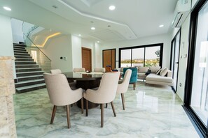 A modern living room with a glass dining table, beige sofas, and a staircase leading to an upper level. - 3-Bedroom Villa with Private Pool & Studio (Bir Bou Rekba)