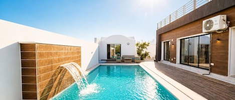A swimming pool with a waterfall feature, a sunlit outdoor seating area, and a modern building facade.
