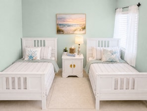 A bedroom with two white beds, a nightstand with a lamp, a painting of a beach scene, and a window with curtains.