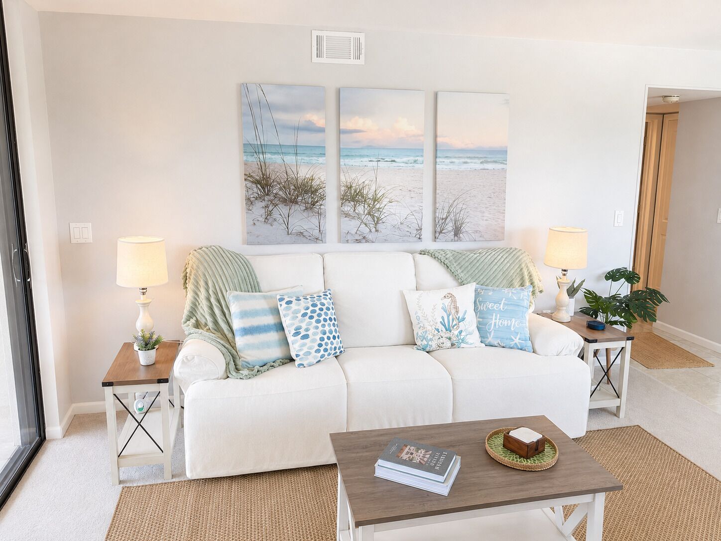 A living room with a white sofa, two side tables with lamps, a coffee table with a book and a decorative item, and a beach-themed wall art above the sofa.