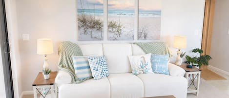 A living room with a white sofa, two side tables with lamps, a coffee table with a book and a decorative item, and a beach-themed wall art above the sofa.