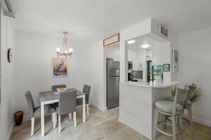 A modern kitchen with a marble island, a refrigerator, and a dining area with a table and chairs. - Sandy Cove Sanctuary | Renovated Ground-Floor (Siesta Key)