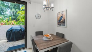 A dining area with a wooden table, chairs, a bowl of fruit, a wall clock, a chandelier, and a painting of a bird. - Sandy Cove Sanctuary | Renovated Ground-Floor Condo, Walk to Village, Private Beach & Fishing Pier (Siesta Key)