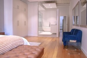 Ett modernt sovrum med en stor säng, en stoppad soffa, ett badrum med glasväggar och en blå fåtölj.