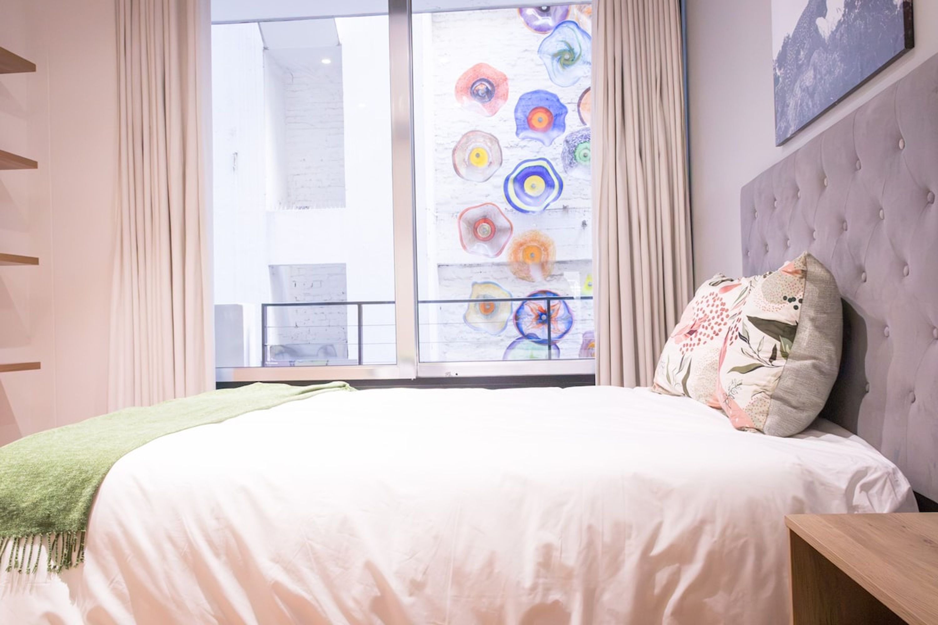 Quarto com uma cama grande, uma janela com arte colorida e vista para um edifício.