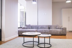 Ein modernes Wohnzimmer mit einem grauen Sofa, zwei runden Tischen und einem großen Fenster, das natürliches Licht hereinlässt.