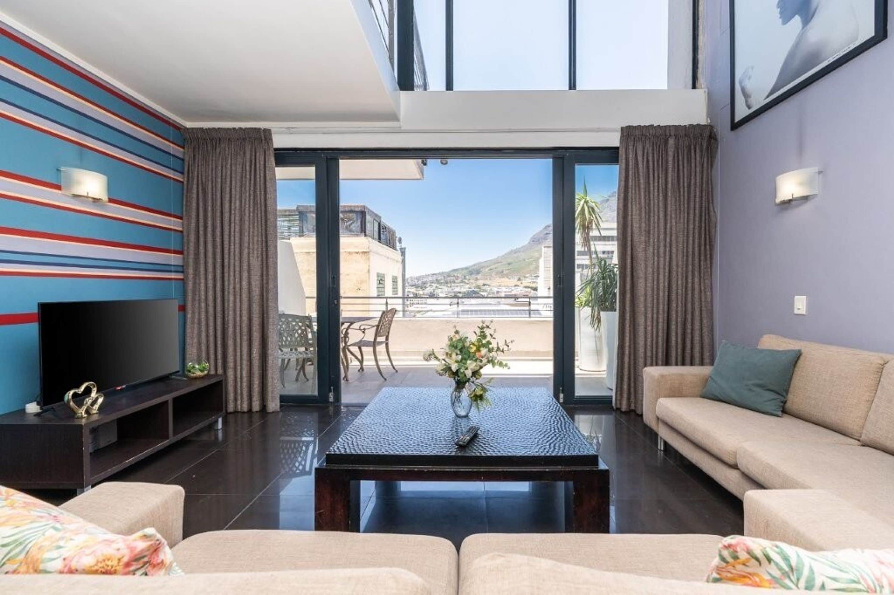 Una sala moderna con un sofá, una mesita de centro y un televisor de pantalla plana. Hay un balcón con vistas a la ciudad y una montaña al fondo.