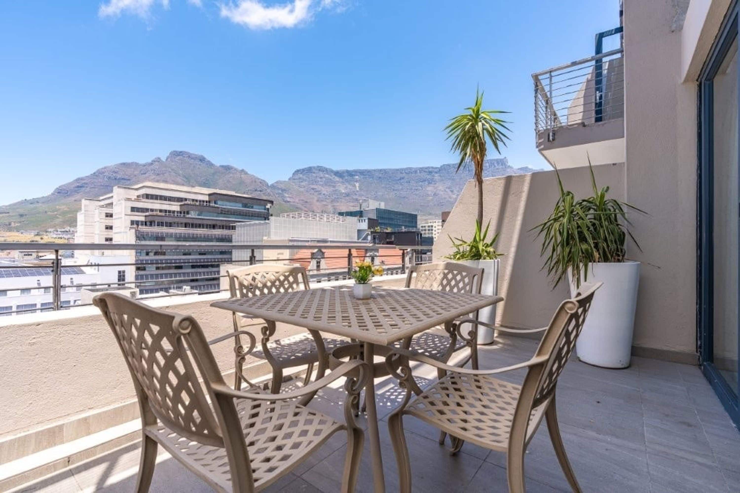 Uma varanda com mesa e cadeiras, plantas em vasos e vista para edifícios e montanhas.
