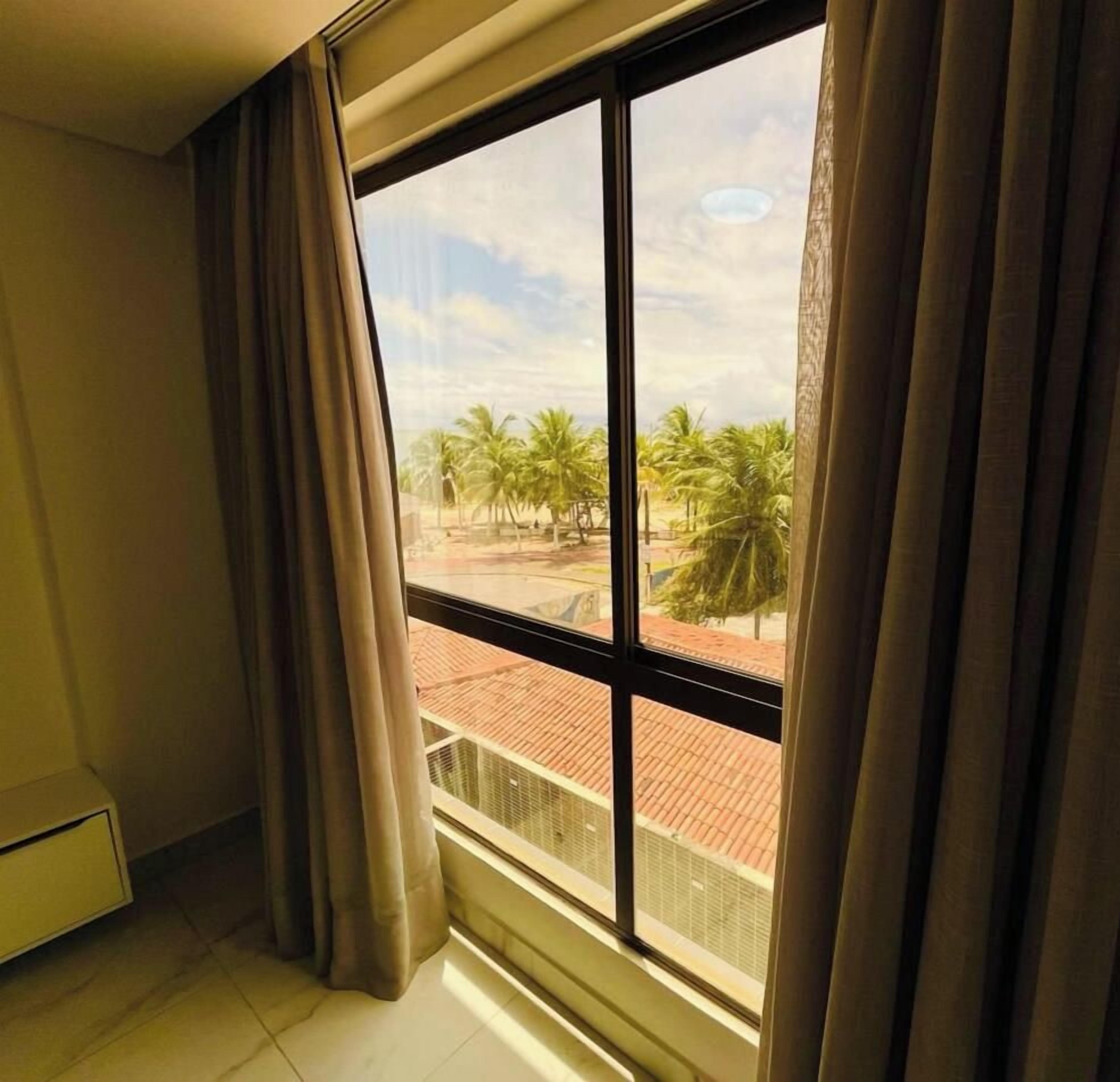 A room with a large window, curtains, and a view of palm trees and a red-roofed building outside.