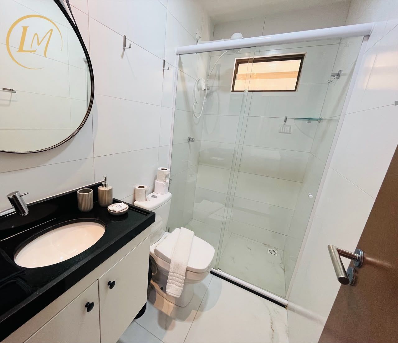 Un baño con ducha envuelta en vidrio, lavamanos con encimera negra y un inodoro con tapa blanca.