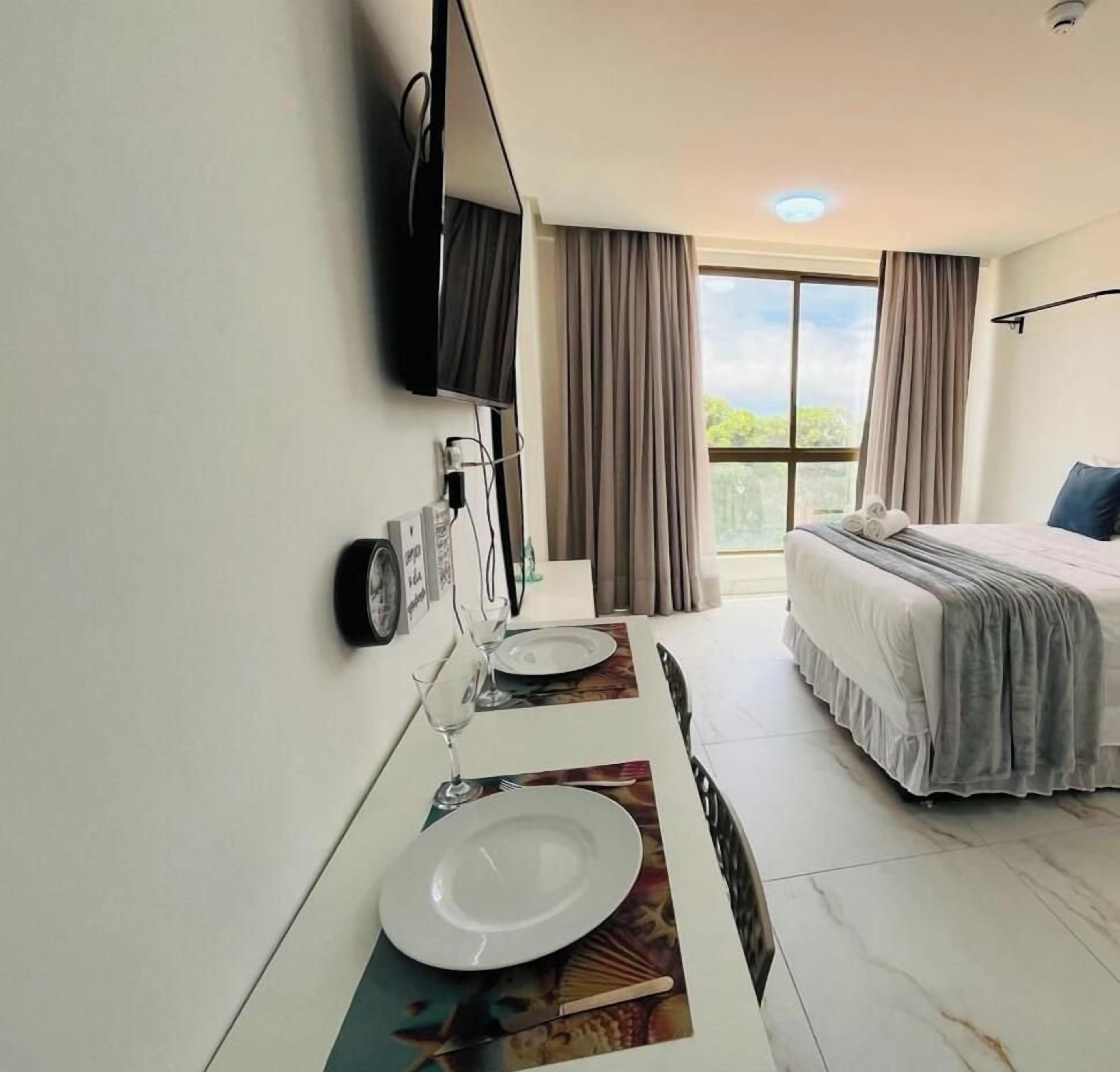 Una habitación de hotel con cama, televisión montada en la pared, una mesa de comedor con plato y vaso, y una ventana con cortinas.